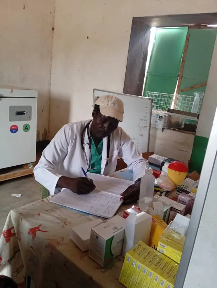 A clinician in a white coat writing in a patient register beside stocked medicines