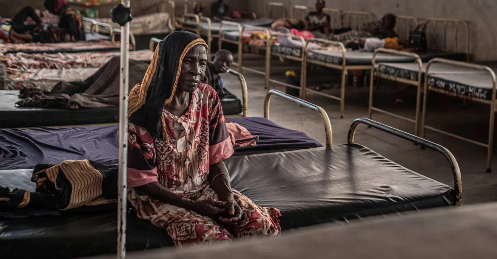 A woman sits on a bed in the hospital's inpatient ward, with other patients resting in the beds behind her